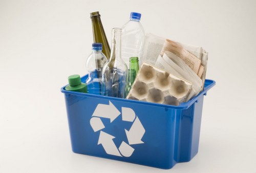 A blue recycling bin filled with various discarded items, including several clear plastic bottles with blue and green caps, an empty glass wine or spirit bottle, a paper carton with egg compartments, and a crumpled newspaper. The bin features a white recycling symbol on the front and is positioned against a plain, light-colored background. The contents are arranged to show the range of different materials, such as plastic, paper, and glass, prepared for rubbish removal or recycling services, typical of garden or household clearance in Erith or surrounding areas. The overall scene emphasizes environmentally conscious waste disposal practices with a focus on recyclable waste as part of rubbish clearance efforts by Garden Clearance Erith.