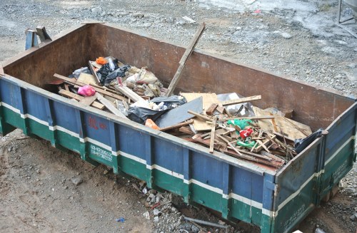 A large, rusted metal skip filled with mixed construction debris and household waste, including wooden planks, plastic bags, and miscellaneous refuse, situated outdoors on a gravel surface. The skip's walls are weathered and stained, with visible signs of rust and age. Some debris spills over the top edges, which are reinforced with metal strips. Surrounding the skip is a rough, uneven ground composed of gravel and dirt, suggesting a construction or clearance site. The scene is outdoors, with natural light illuminating the waste materials and casting minimal shadows, indicative of overcast weather or diffused sunlight. This image reflects the typical clearing of rubbish associated with garden and property clearance services provided by Garden Clearance Erith, with a focus on removing various types of waste material from residential or commercial sites in Erith and nearby areas.