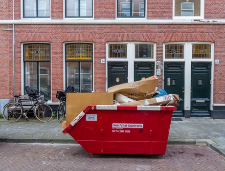 A bright red skip bin positioned on the sidewalk in front of a row of brick terrace houses in Erith, with its side panel displaying a local waste management company number. The bin is filled with a variety of rubbish items, including large cardboard boxes, paper, and plastic packaging, with some debris protruding from the top. To the left of the bin, two bicycles are parked against the building's brick façade, which features tall, narrow windows with metal bars and white frames. The doors of the houses are painted dark green, each with a brass door handle and letterbox, and all have small clear glass panes above the doors. The scene is illuminated evenly, indicating daytime overcast weather, with the environment suggesting a typical residential area requiring rubbish removal services from Garden Clearance Erith, located in the local postcode region.