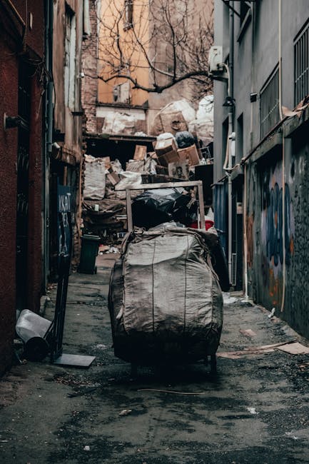 A street scene showing a white waste collection truck with a rusted rear compartment, positioned alongside a row of older multi-story buildings with varied brick and stucco facades. The truck's rear l